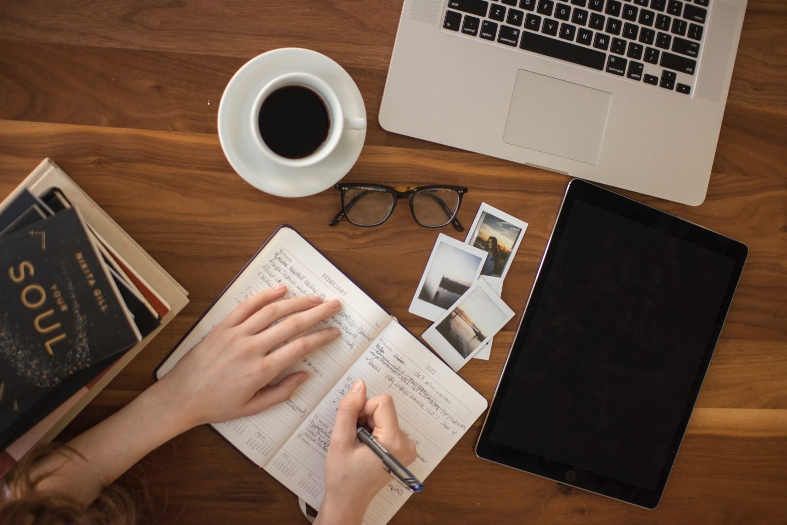 A woman's hands writing on a notebook with a cup of coffee and a laptop on a wooden table.